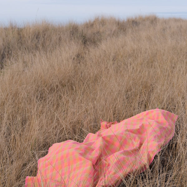 Person wrapped in a pink blanket lying in dry grass with a clear sky. laid towel with tassels. Checker Gingham Picnic Blanket.